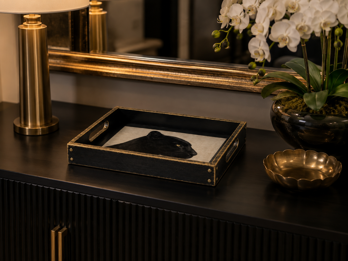 A black rectangular decorative tray with gold trim and handles, styled on a dark console table next to a gold lamp, white orchids, and a small gold bowl.