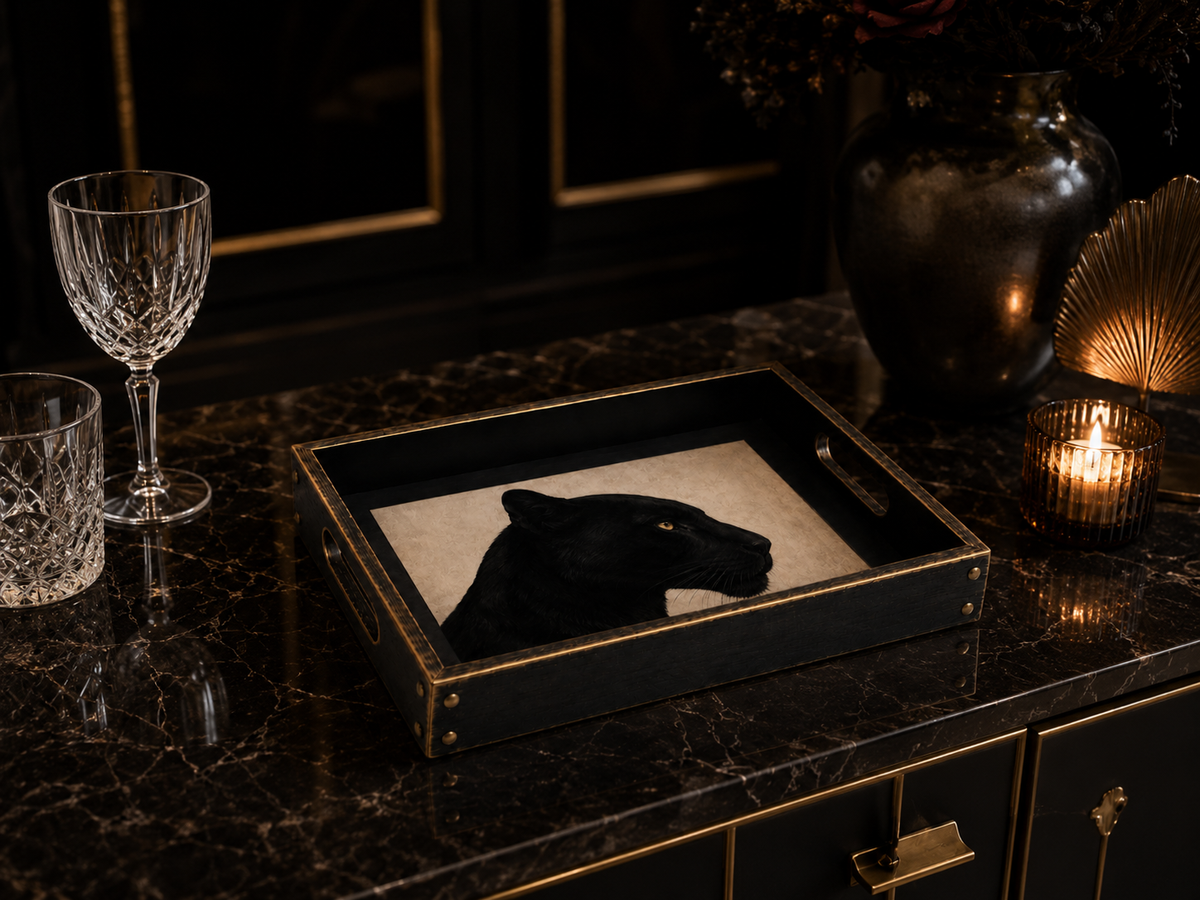 A rectangular black tray with gold trim and handles, featuring a black panther head silhouette on a cream insert, styled on a dark marble sideboard with a wine glass, candle, and vase nearby.