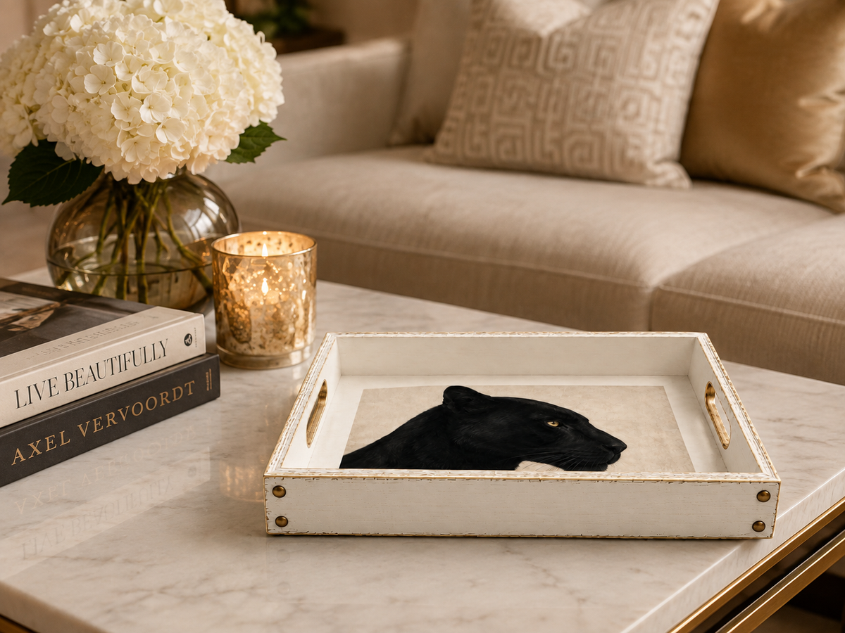 A white rectangular decorative tray with gold-toned handles and corner accents sits on a marble coffee table in a styled living room. The tray contains a black panther head mounted in the center.