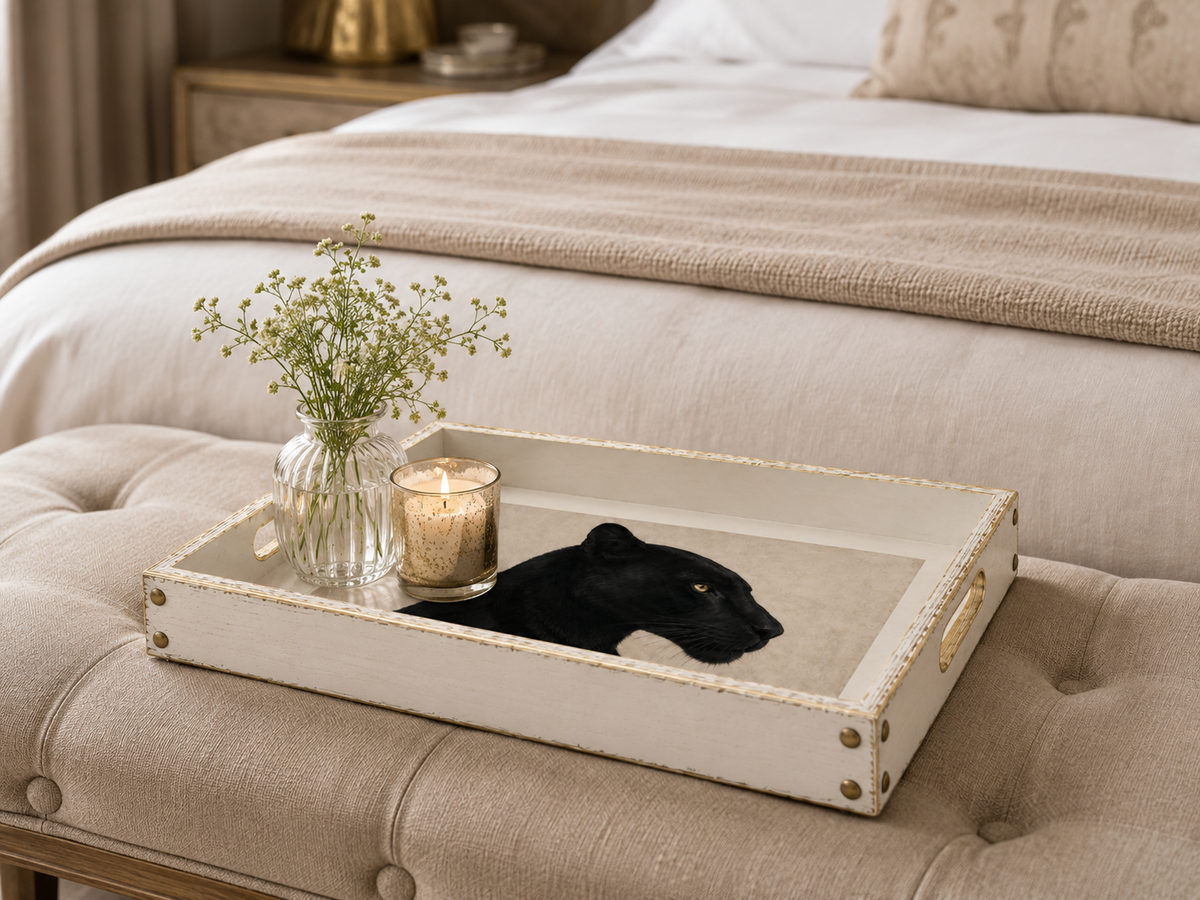 A rectangular white tray with gold trim and handles sits on a tufted bench in a bedroom setting. The tray holds a small glass vase with baby's breath and a lit candle, with a black panther image inside the tray.