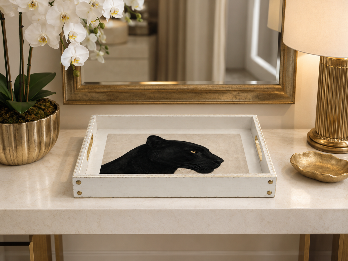 A black panther head image on a beige linen background, presented inside a white wooden serving tray with brass corner rivets, styled on a marble console table.
