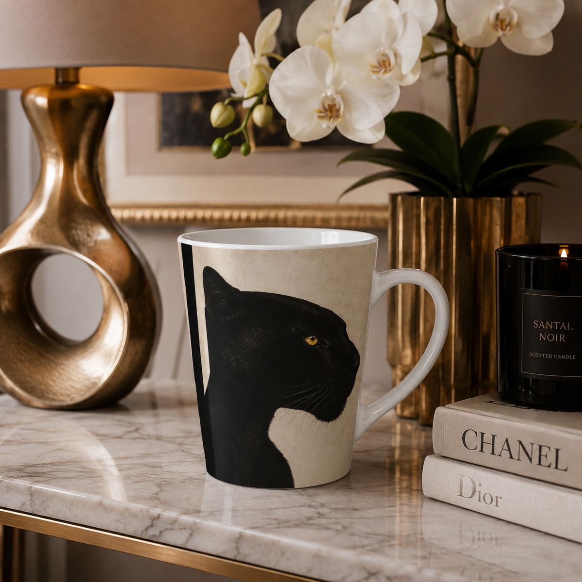 A ceramic mug featuring a black panther illustration on a cream background, styled on a marble side table next to a gold lamp, white orchids, fashion designers book stack, and a Santal Noir candle.