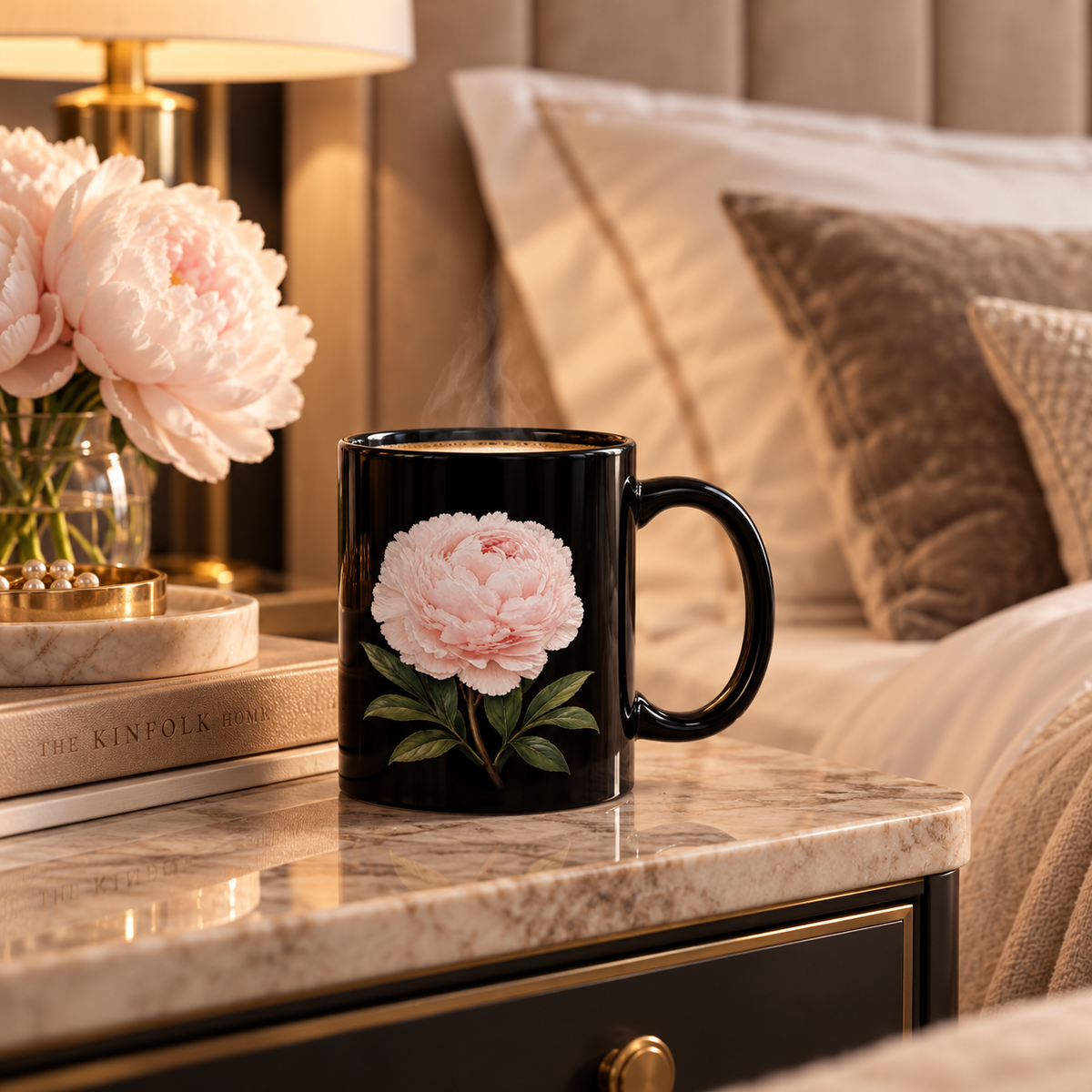 A black ceramic mug with a large pink peony and green leaves printed on the side, set on a marble bedside table in a cozy bedroom scene.