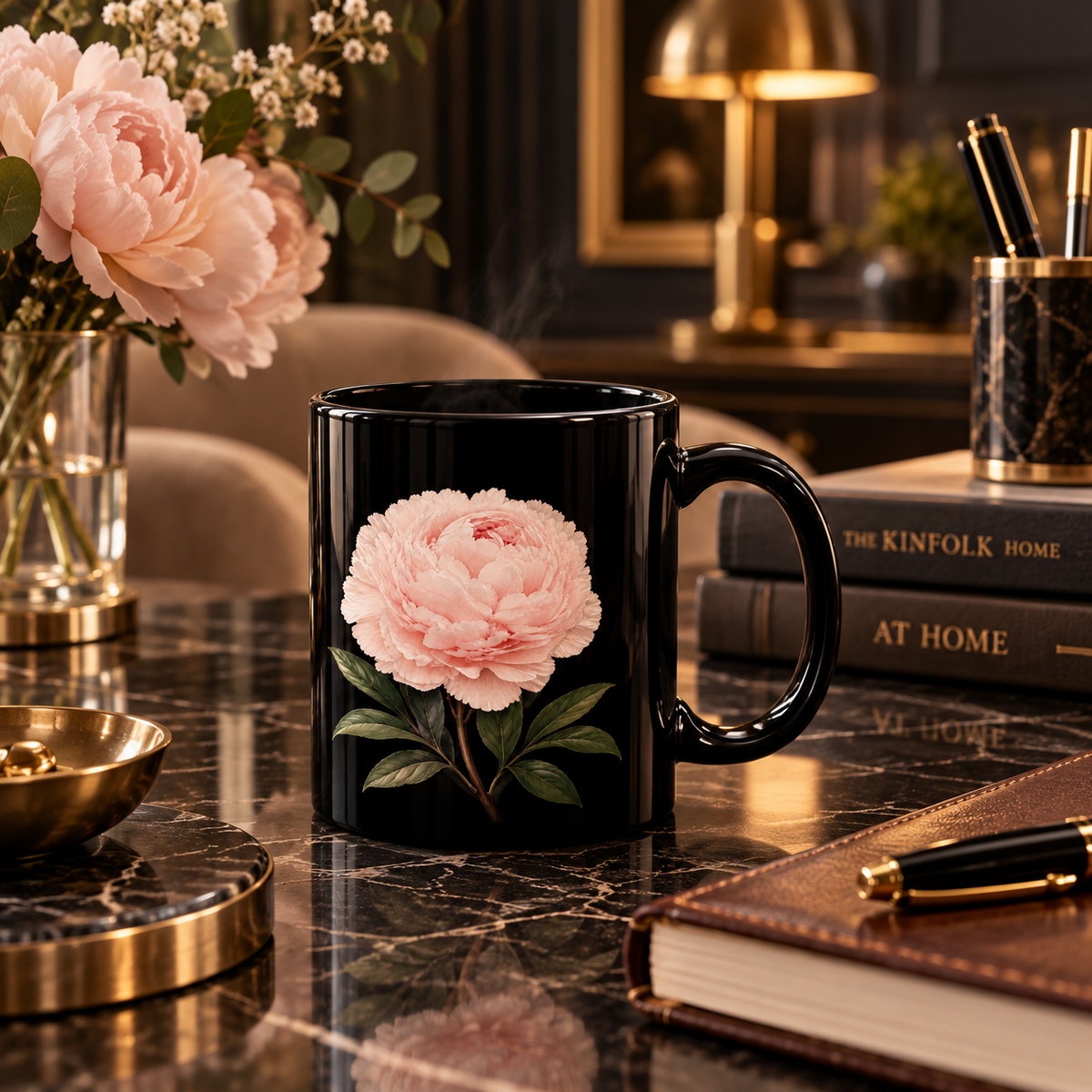 A glossy black ceramic mug featuring a large pink peony flower with green leaves, set on a dark marble desk in a cozy, warmly lit room with books, a lamp, and flowers in the background.