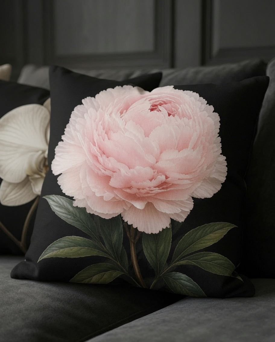 A square black throw pillow featuring a large, realistic light pink peony flower with green leaves, styled on a dark gray sofa.