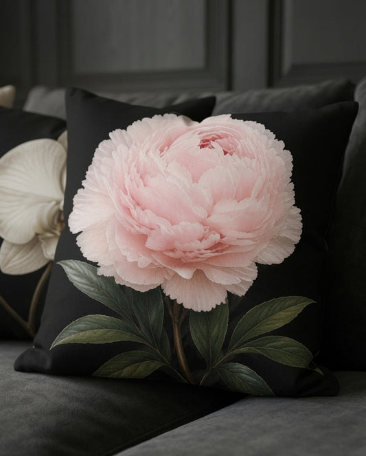 A square black throw pillow featuring a large, realistic light pink peony flower with green leaves, styled on a dark gray sofa.
