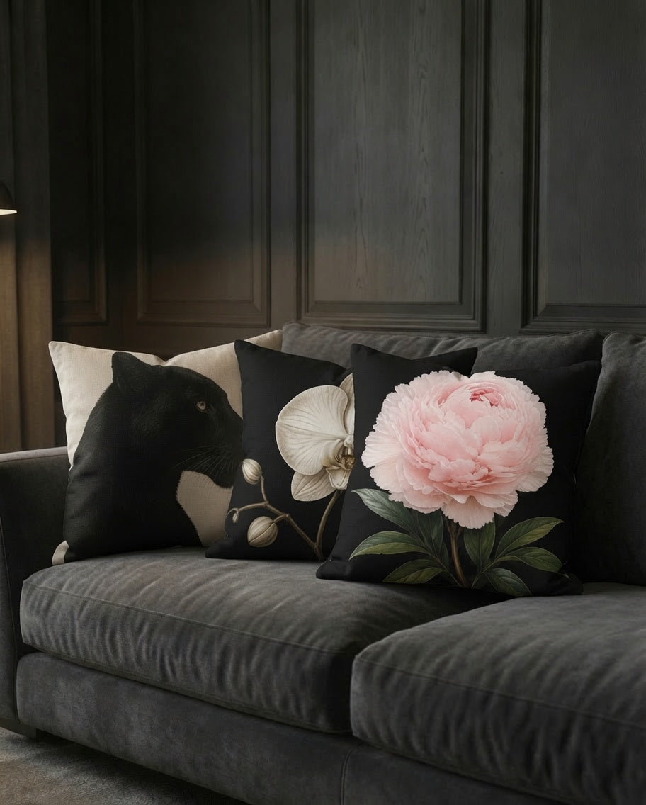 Three decorative throw pillows styled on a dark gray velvet sofa against a dark wood-paneled wall. The pillows include a black pillow with a white panther design, a black pillow with a white orchid, and a black pillow with a large pink peony flower and green leaves.