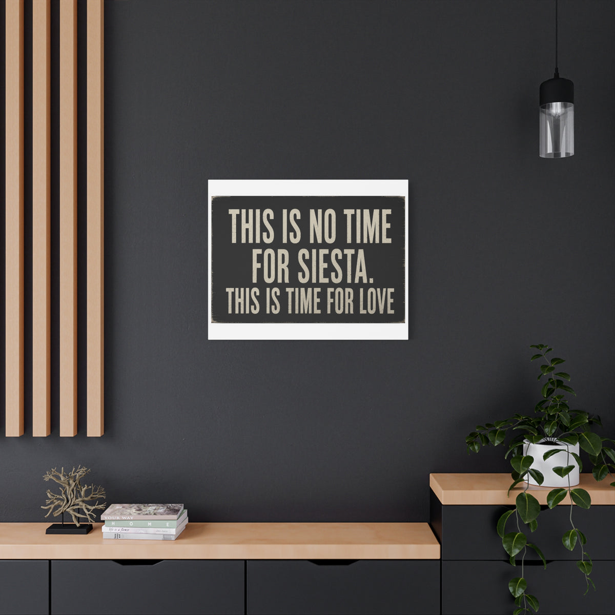 A square wall art print with the text 'THIS IS NO TIME FOR SIESTA. THIS IS TIME FOR LOVE' in beige lettering on a dark charcoal background, displayed on a dark gray wall in a modern room setting with a wooden console, potted plant, and hanging light.