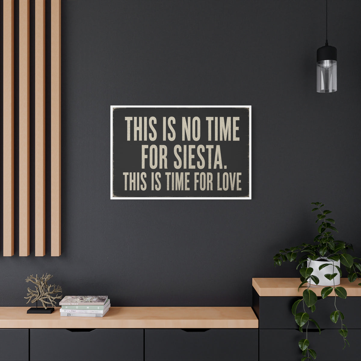 A square wall art print with the text 'THIS IS NO TIME FOR SIESTA. THIS IS TIME FOR LOVE' in beige lettering on a dark charcoal background, displayed on a dark gray wall in a modern room setting with a wooden console, potted plant, and hanging light.