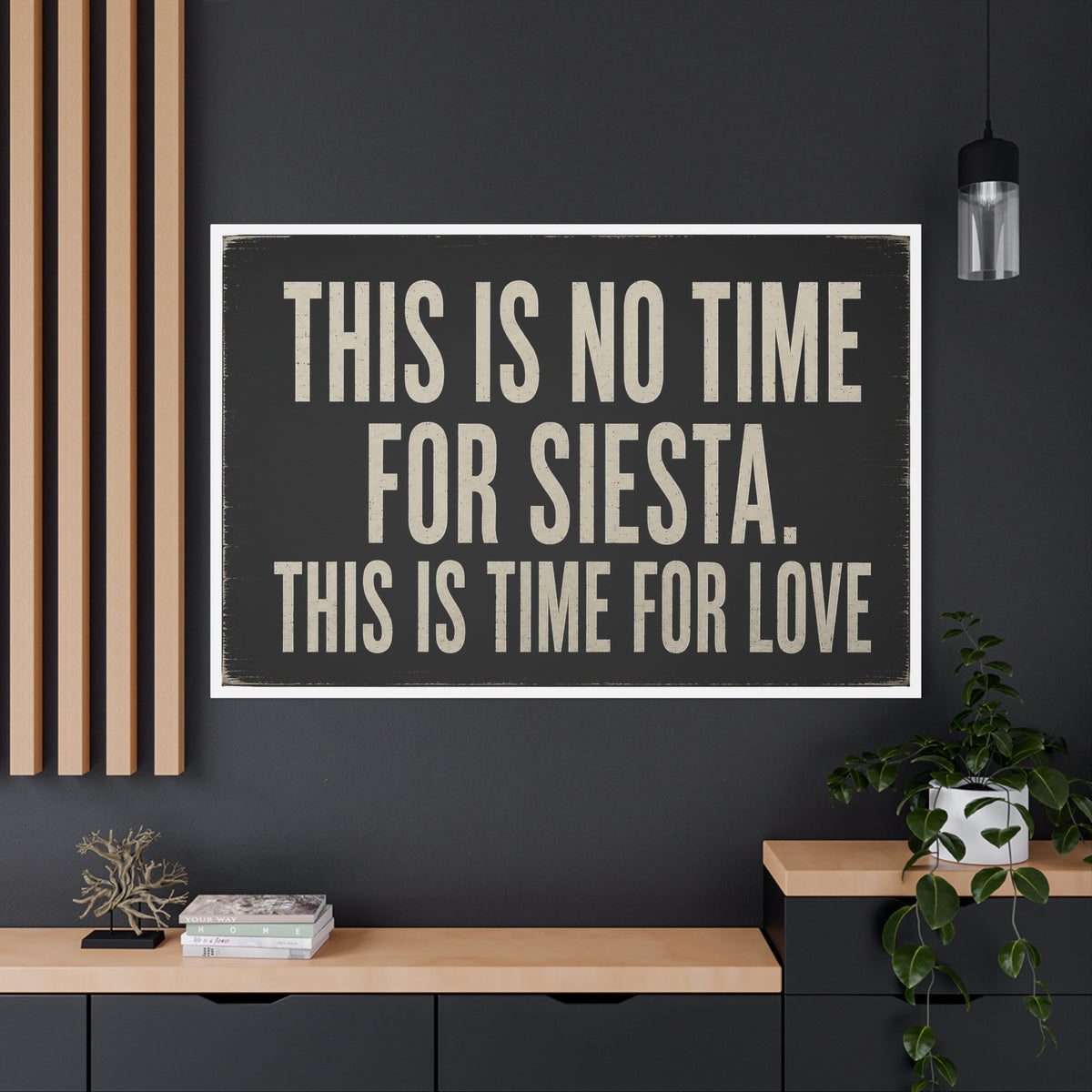 Extra large canvas with the text 'THIS IS NO TIME FOR SIESTA, THIS IS TIME FOR LOVE' in beige block letters on a black background on a grey living room wall.