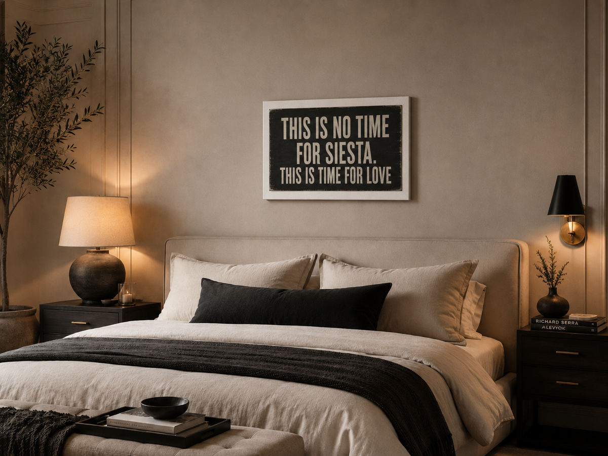 A framed typographic wall art print hangs above a neatly made bed in a warm, modern bedroom. The print reads, 'THIS IS NO TIME FOR SIESTA. THIS IS TIME FOR LOVE'.