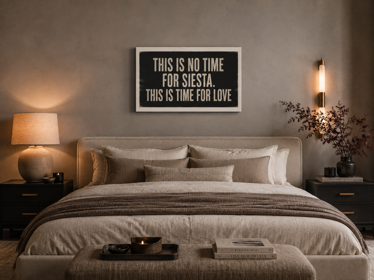 A framed wall art print with the text 'THIS IS NO TIME FOR SIESTA. THIS IS TIME FOR LOVE' in cream lettering on a dark background, displayed above a neatly made bed in a warm, modern bedroom setting.
