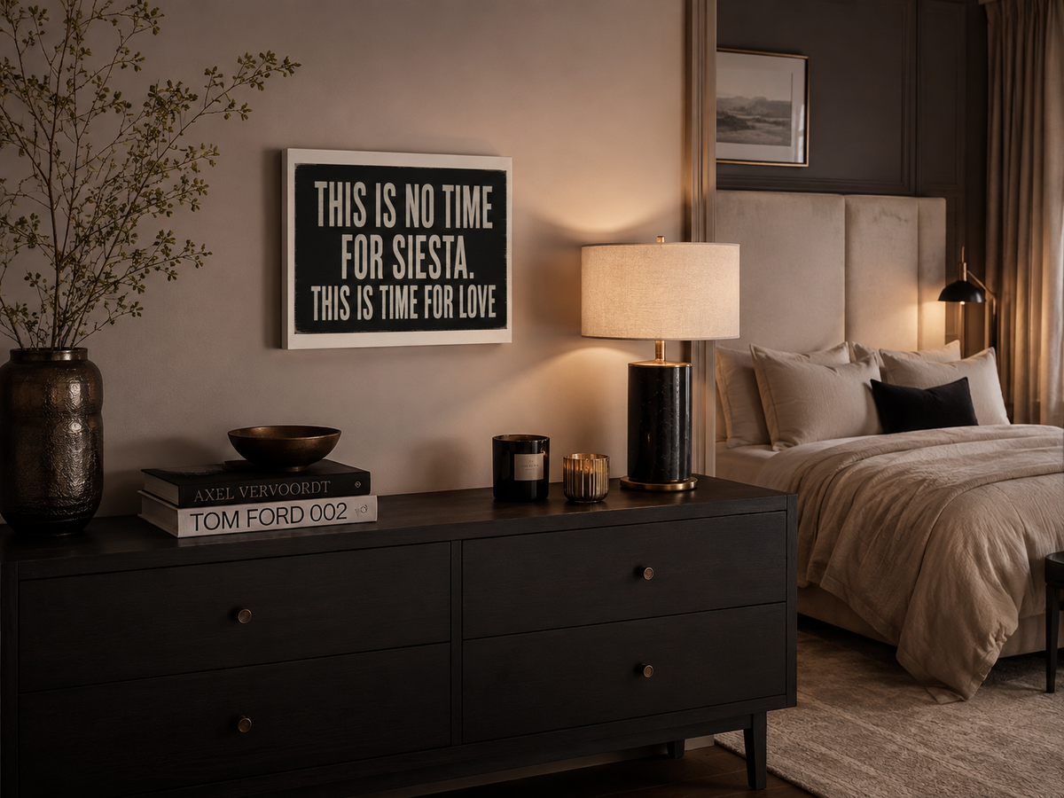 A framed typographic wall art print hangs above a dark wood dresser in a styled bedroom. The print reads, 'THIS IS NO TIME FOR SIESTA. THIS IS TIME FOR LOVE'.