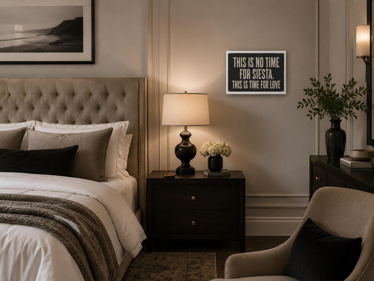 A framed typographic wall art print hanging on a beige wall in a styled bedroom. The print reads, 'THIS IS NO TIME FOR SIESTA, THIS IS TIME FOR LOVE' in white text on a dark charcoal background.