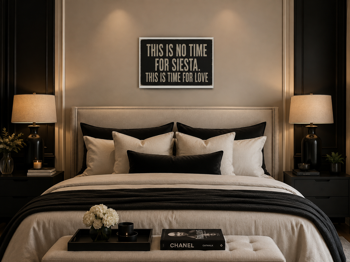 A styled bedroom scene with a neatly made bed, black and white bedding, matching pillows, bedside tables with lamps, and a framed quote on the wall above the headboard. The quote reads, 'THIS IS NO TIME FOR SIESTA. THIS IS TIME FOR LOVE'.
