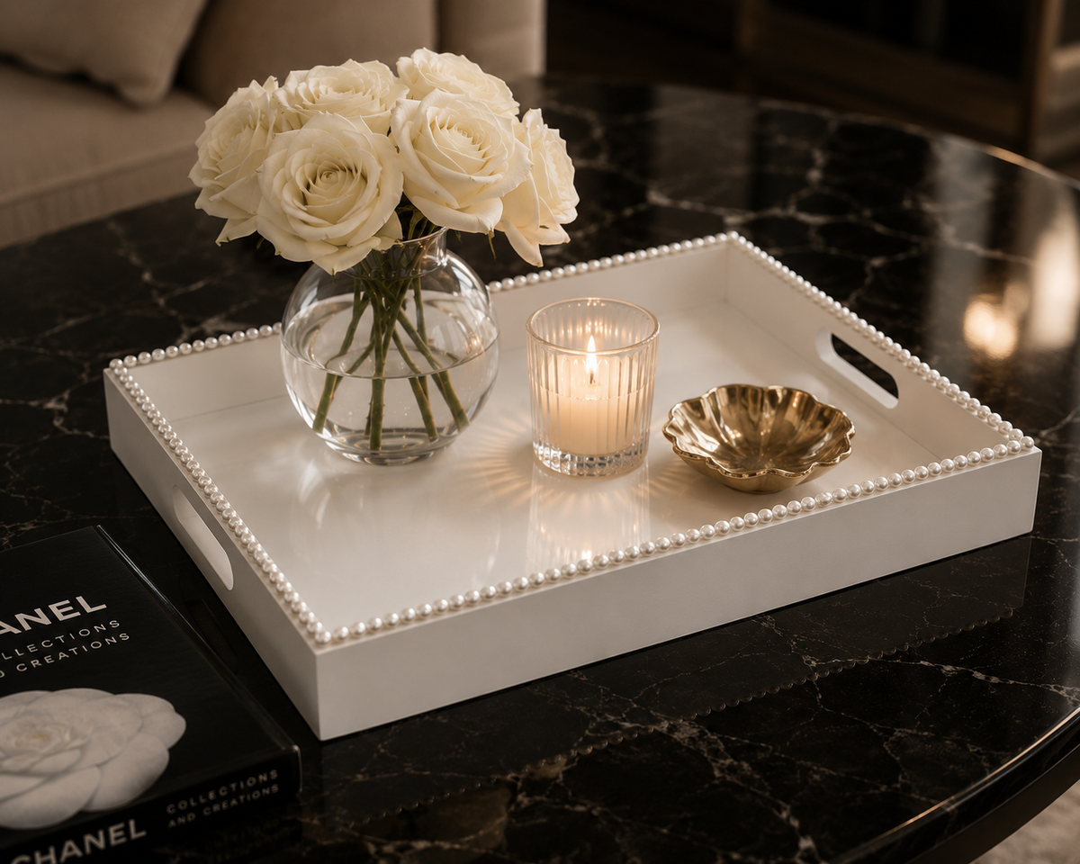 A rectangular white decorative tray with a beaded edge and cutout handles sits on a black marble table. The tray is styled with a clear glass vase of white roses, a lit candle, and a small gold dish.