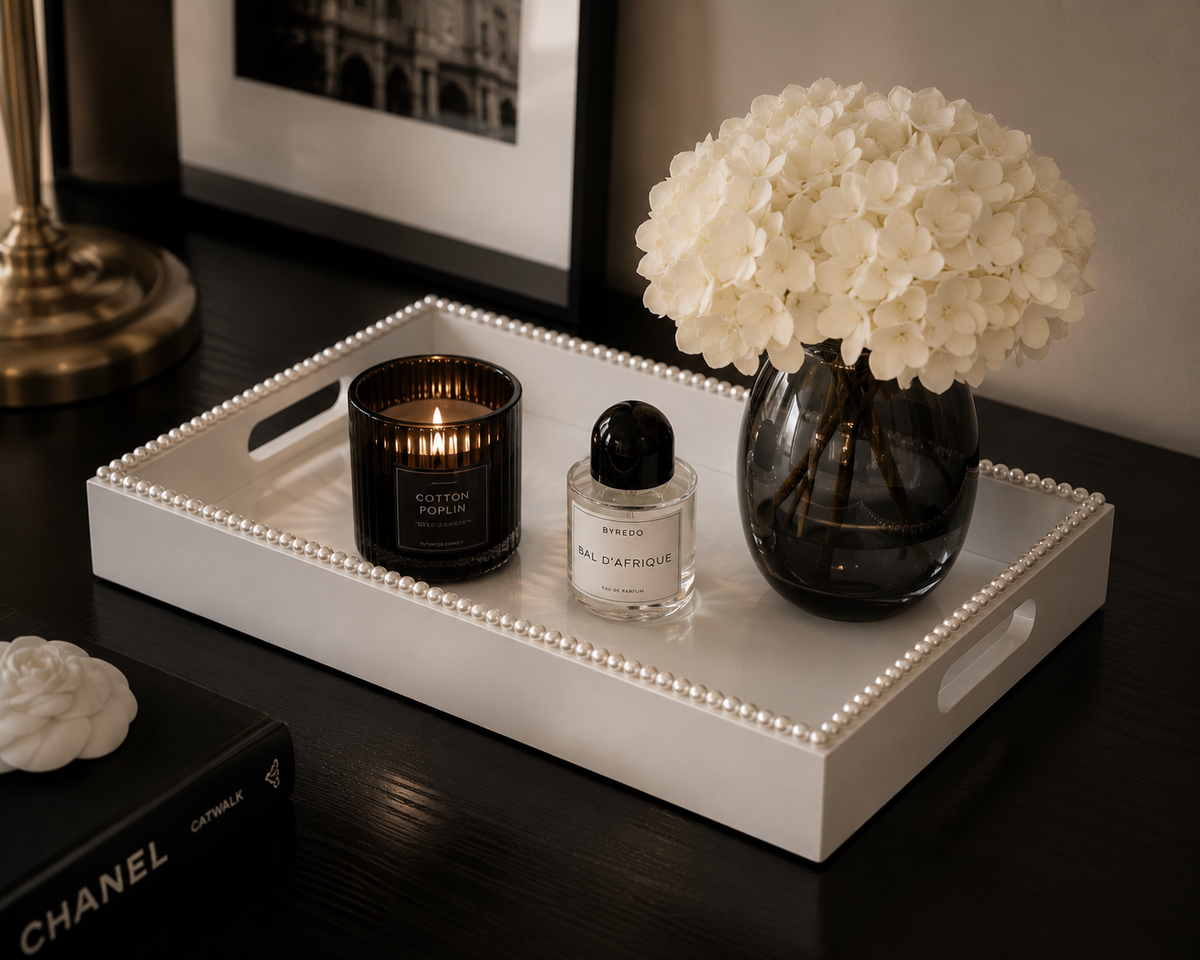 A white decorative tray with a beaded edge sits on a dark wooden table, styled with a lit black candle, a small clear perfume bottle, and a dark glass vase filled with white flowers. A fashion design book is partially visible in the foreground.