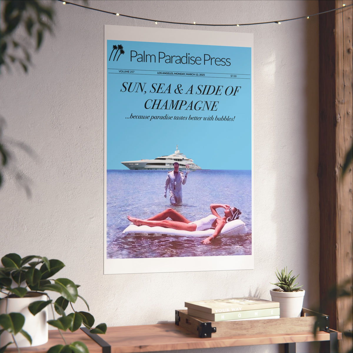 A framed or mounted poster reading 'Palm Paradise Press' hangs on a light wall above a wooden console. The poster shows a bright blue sky and ocean with a yacht and a woman lounging on a float, with the headline 'Sun, Sea & a Side of Champagne.