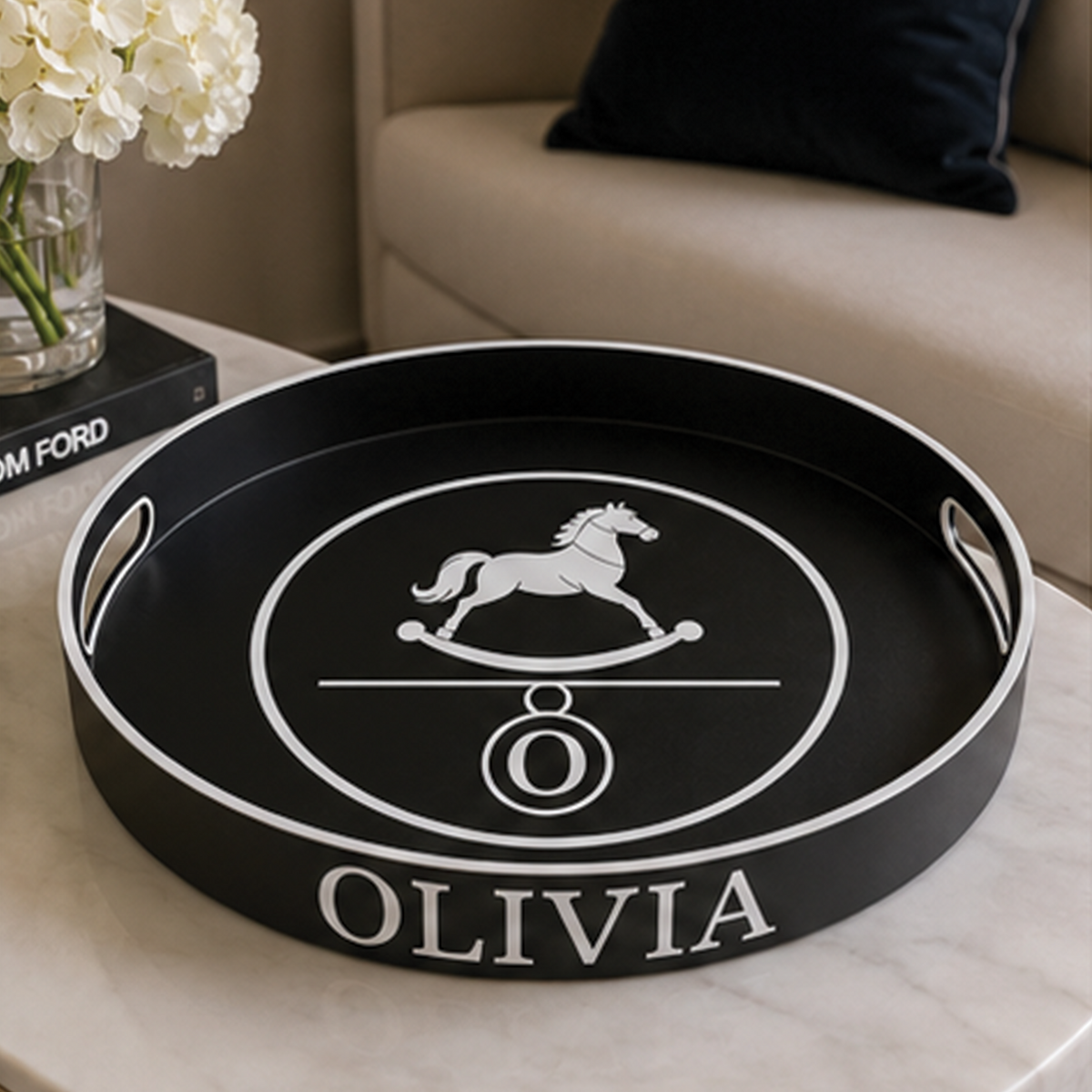 A round black serving tray with white trim and a white rocking horse logo, personalized with the name 'OLIVIA' on the front, displayed on a marble table in a living room setting.