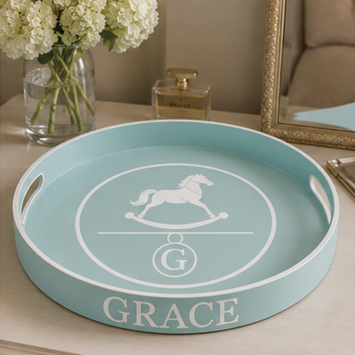 A round light blue serving tray with white handles and a white rocking horse monogram design, personalized with the name 'GRACE' at the bottom, displayed on a wooden table in a styled home setting.