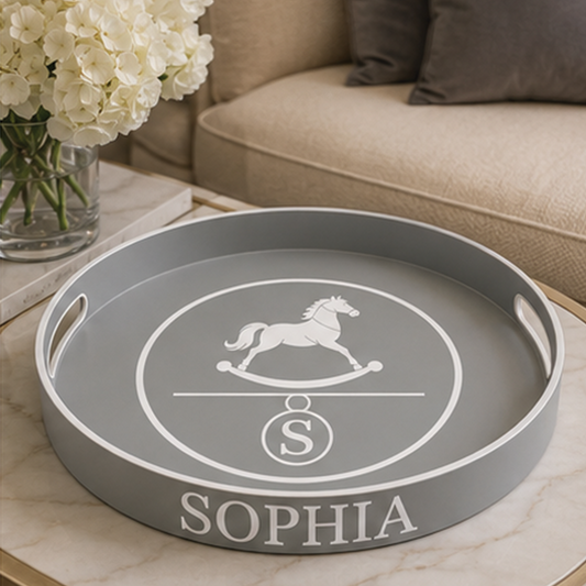 A round gray decorative tray with white printed graphics and the name 'SOPHIA' on the front, placed on a light marble surface in a living room setting with a sofa and flowers in the background.