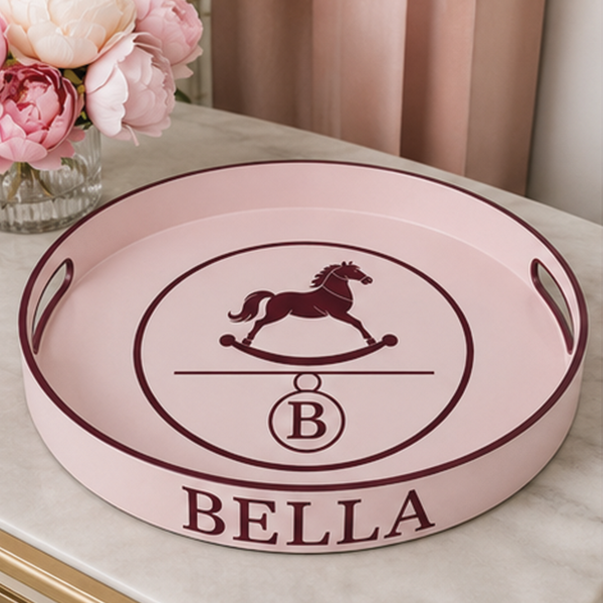 A round pink serving tray with dark burgundy accents, featuring a rocking horse emblem, a monogrammed 'B', and the name 'BELLA' printed on the front. The tray is styled on a marble surface next to a vase of pink flowers.