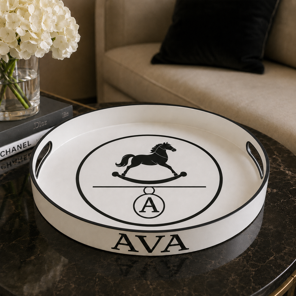 A round white serving tray with black trim and handles sits on a dark marble table in a living room setting. The tray features a black rocking horse logo inside a circle with the initials 'AVA' printed at the bottom.